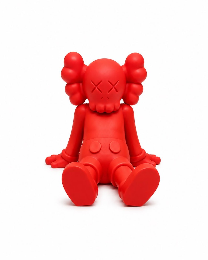 Op de afbeelding: Een effen rode vinyl figuur in een zittende positie. De figuur heeft grote, afgeronde oren, doorgestreepte ogen en een kleine mond. De figuur draagt een korte broek en heeft zijn handen op zijn benen. Een verzamelobject.