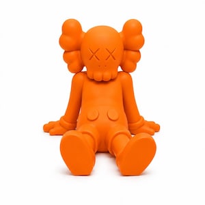 Op de afbeelding: Fel oranje vinyl figuur in een zittende positie. Het figuur heeft een afgeronde kop met doorgestreepte ogen en een schedelachtige mond. De armen en benen zijn uitgestrekt en het heeft grote, afgeronde voeten. Het figuur is effen oranje van kleur.