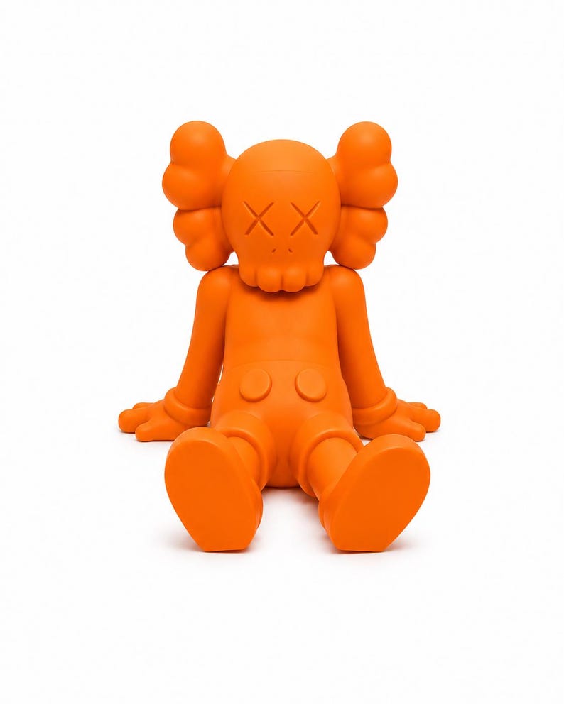 Op de afbeelding: Fel oranje vinyl figuur in een zittende positie. Het figuur heeft een afgeronde kop met doorgestreepte ogen en een schedelachtige mond. De armen en benen zijn uitgestrekt en het heeft grote, afgeronde voeten. Het figuur is effen oranje van kleur.