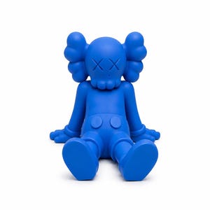 Op de afbeelding: Een effen blauw beeldje van een zittend stripfiguur met doorgestreepte ogen. De figuur heeft grote, afgeronde oren, een schedelachtig gezicht en draagt een korte broek. De armen en benen zijn uitgestrekt en de figuur zit op een witte achtergrond.