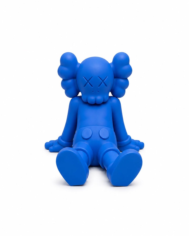 Op de afbeelding: Een effen blauw beeldje van een zittend stripfiguur met doorgestreepte ogen. De figuur heeft grote, afgeronde oren, een schedelachtig gezicht en draagt een korte broek. De armen en benen zijn uitgestrekt en de figuur zit op een witte achtergrond.