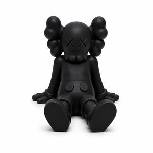 Op de afbeelding: Een zwarte vinyl figuur van een zittend stripfiguur met doorgestreepte ogen. De figuur heeft grote, afgeronde oren, een schedelachtig gezicht en gehandschoende handen. De figuur zit met uitgestrekte benen en voeten plat op de grond.