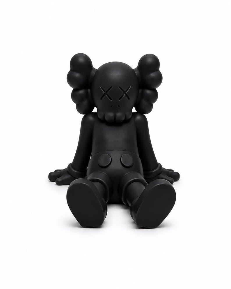 Op de afbeelding: Een zwarte vinyl figuur van een zittend stripfiguur met doorgestreepte ogen. De figuur heeft grote, afgeronde oren, een schedelachtig gezicht en gehandschoende handen. De figuur zit met uitgestrekte benen en voeten plat op de grond.
