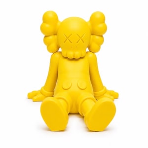 Op de afbeelding: Een heldergele vinyl figuur in een zittende positie. De figuur heeft een ronde kop met gekruiste ogen, grote oorvormige vormen en een kleine mond. De armen zijn uitgestrekt en de benen gebogen.