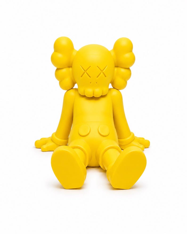 Op de afbeelding: Een heldergele vinyl figuur in een zittende positie. De figuur heeft een ronde kop met gekruiste ogen, grote oorvormige vormen en een kleine mond. De armen zijn uitgestrekt en de benen gebogen.