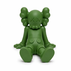 Op de afbeelding: Een groene vinyl figuur van een zittend stripfiguur met doorgestreepte ogen. De figuur heeft grote, afgeronde oren, gehandschoende handen en een korte broek. De figuur zit met uitgestrekte benen en platte voeten op de grond.