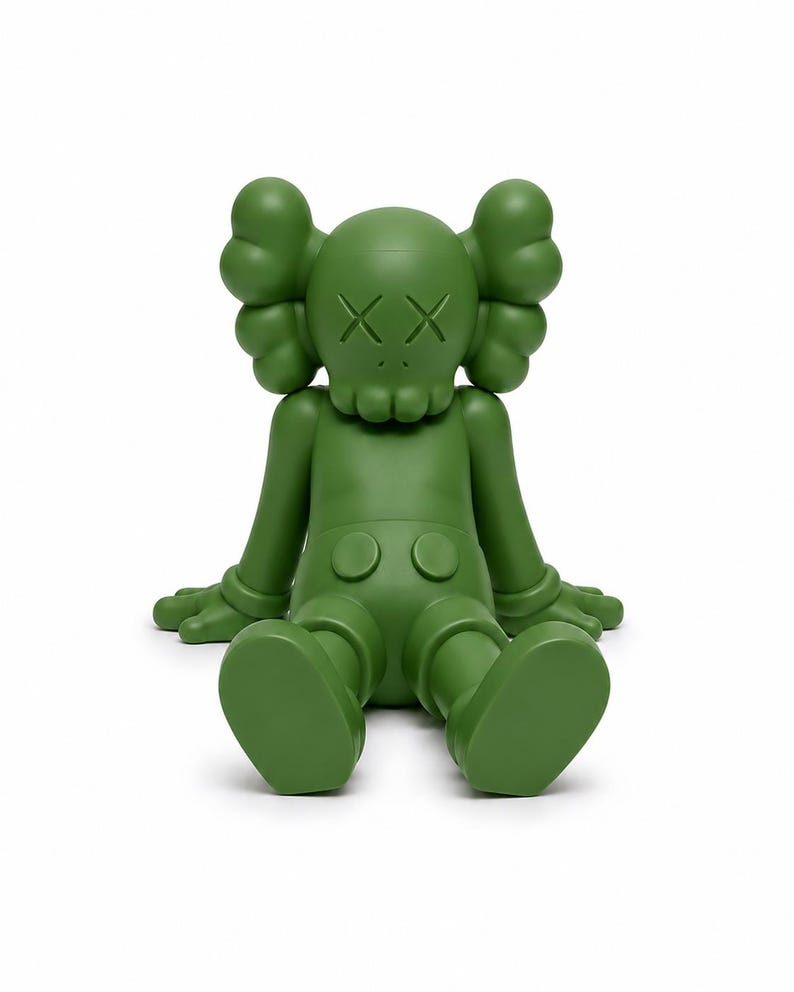 Op de afbeelding: Een groene vinyl figuur van een zittend stripfiguur met doorgestreepte ogen. De figuur heeft grote, afgeronde oren, gehandschoende handen en een korte broek. De figuur zit met uitgestrekte benen en platte voeten op de grond.