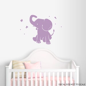 May include: A lavender-colored elephant decal with a whimsical design, featuring a sitting elephant with its trunk raised. Butterflies and dots are scattered around the elephant. The decal is shown above a white crib.