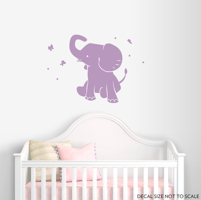 May include: A lavender-colored elephant decal with a whimsical design, featuring a sitting elephant with its trunk raised. Butterflies and dots are scattered around the elephant. The decal is shown above a white crib.