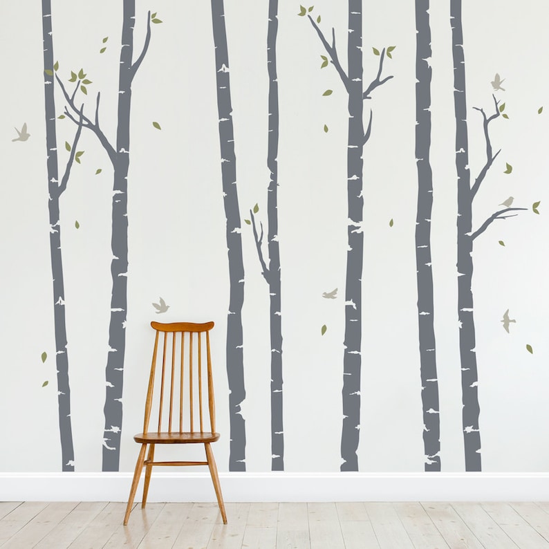 May include: A wall decal featuring a gray birch tree forest with small green leaves and gray birds. The decal is applied to a white wall with a wooden floor and a wooden chair in the foreground.