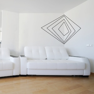 May include: A modern living room featuring a white sofa and armchair set. The sofa has a two-cushion design with a tufted back. A black geometric design adorns the white wall. The room has light wood flooring.