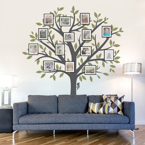 May include: A gray tree silhouette with green leaves and 16 black picture frames on a white wall. The frames are filled with family photos. A gray couch with pillows sits in front of the wall.