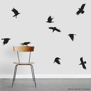 May include: A set of nine black crow silhouette wall decals. The decals are arranged in a random pattern on a white wall. A wooden chair is in the foreground.
