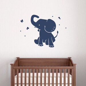 May include: A navy blue elephant wall decal with a whimsical design. The elephant is sitting with its trunk raised, surrounded by small butterflies and dots. The decal is placed on a white wall above a wooden crib.