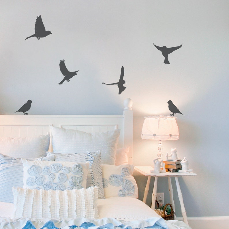 May include: A white bed with a white headboard and a white lamp on a white nightstand. The bed is made with white and blue bedding and has a blue and white throw pillow. There are five black bird silhouettes on the wall behind the bed.