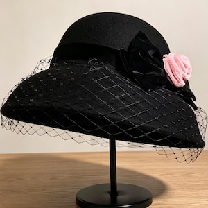 May include: A black felt hat with a wide brim and a black ribbon band. The hat is adorned with a large black fabric flower and a smaller light pink fabric rose. A black netting veil with a diamond pattern covers the front of the brim.
