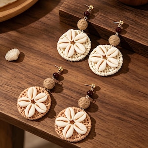 Op de afbeelding: Close-up van drie paar oorbellen. Elke oorbel heeft een geweven, cirkelvormige basis versierd met witte schelpen in een bloemmotief. De oorbellen hebben goudkleurige pinnen en zijn geaccentueerd met bruine kralen en geweven bollen.