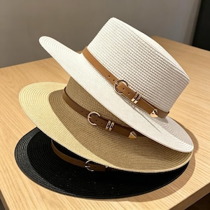 Könnte beinhalten: Ein Stapel aus drei Fedora-Hüten in Weiß, Beige und Schwarz. Jeder Hut hat eine geflochtene Strohstruktur, eine flache Krempe und ein braunes Kunstlederband mit einer goldfarbenen Metallschnalle und einem kleinen herzförmigen Anhänger.