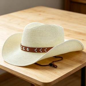 May include: A cream-colored straw cowboy hat featuring a brown faux leather band adorned with silver-toned metal studs and chevron-shaped accents. The hat includes a dark brown chin cord with an adjustable toggle for a secure fit.