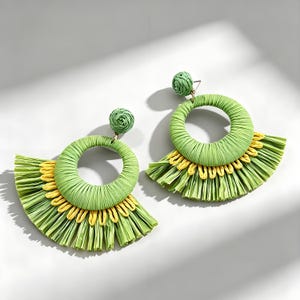 Puede incluir: Un par de pendientes colgantes con un aro circular de rafia trenzada verde unido a un pequeño perno trenzado verde. La mitad inferior de cada aro está adornada con un fleco en capas de hebras de rafia de color amarillo brillante y verde claro.