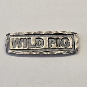 May include: A rectangular metal tag with a hammered, textured border and a dark, recessed background. The words WILD PIG are embossed in raised, metallic capital letters across the center of the piece, creating a distinct contrast in depth.
