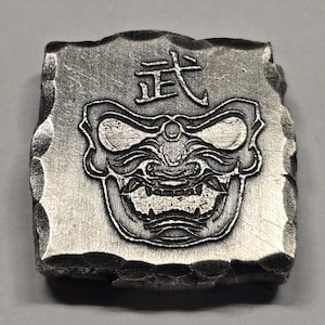 May include: A square, silver-toned metal plaque with a hammered, uneven edge. The center features an embossed, stylized mask with bared teeth and prominent eyes. Above the mask, the Japanese kanji character for martial, 武, is clearly engraved.