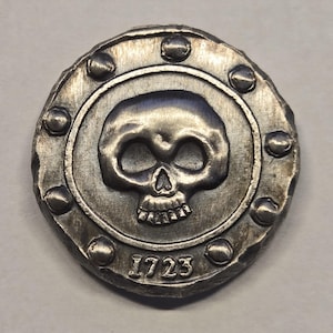 May include: A circular silver-toned metal coin featuring a raised skull design in the center. The outer border is decorated with small, evenly spaced raised studs. The number 1723 is embossed at the bottom of the coin, below the skull graphic.