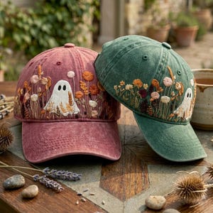 May include: Two baseball caps, one in a faded red and one in a muted green, each featuring detailed embroidery of a small white ghost surrounded by various wildflowers in shades of orange, yellow, and cream, with a structured six-panel design.