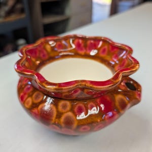 May include: A ceramic wax warmer featuring a scalloped rim and a glossy glaze with a mottled pattern of deep red, brown, and orange tones. The bowl has a smooth, cream-colored interior basin and a small side opening for ventilation.