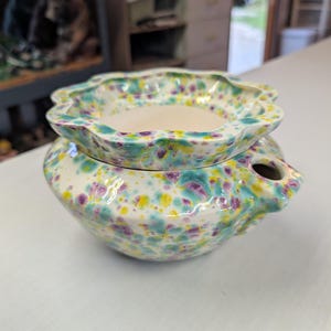 May include: A ceramic wax warmer featuring a two-piece design with a scalloped edge bowl resting on a rounded base. The exterior is finished in a glossy white glaze with a speckled pattern of teal, purple, and yellow spots throughout the surface.