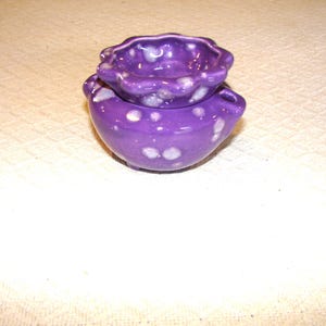 May include: A small, two-piece ceramic container in a vibrant purple hue, speckled with white accents. The top piece has a scalloped edge, and the bottom piece has two small handles. This decorative item is likely for storing small items.