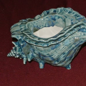 May include: A ceramic seashell-shaped bowl with a turquoise and blue glaze. The bowl has a textured surface and a white interior. It is resting on three small feet and is suitable for decorative purposes.