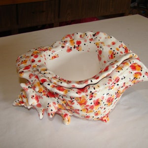 May include: A ceramic seashell-shaped bowl with a white base, adorned with red, orange, and black floral patterns. The bowl has a textured surface and a smooth, white interior. It is a decorative piece.