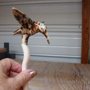 May include: A ceramic hummingbird sculpture with a speckled brown and cream body and wings. The bird's beak is long and curved, and the sculpture is held in a hand. The background is a neutral-toned wall and wooden surface.