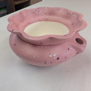 May include: A pink ceramic pot with a speckled design. The pot has a ruffled rim and a small handle. The interior is white. The pot is sitting on a white surface.