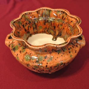 May include: A ceramic planter with a scalloped rim and two side handles. The planter has a speckled orange glaze with green and black accents. The interior is a light cream color. A decorative piece for plants.