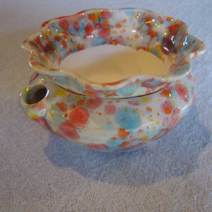 May include: A ceramic oil warmer with a speckled design in red, blue, and yellow. The bowl has a scalloped rim and a small spout. The interior is white.