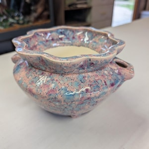 May include: A ceramic oil warmer featuring a scalloped rim and a speckled glaze in shades of pink, blue, and purple. The bowl has a smooth cream-colored interior, a rounded base, and a small side opening for a tea light candle to be placed inside.