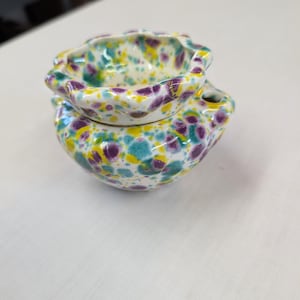 May include: A two-piece ceramic container with a speckled design. The container is white with teal, purple, and yellow spots. The top piece is a small, scalloped bowl that sits on top of a larger, rounded bowl.