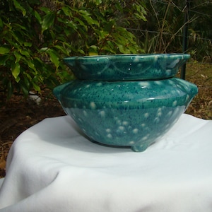 May include: A two-piece teal ceramic bowl set. The larger bowl has a rounded shape with three small feet and a speckled pattern. The smaller bowl sits on top, featuring a scalloped rim. The set is glazed with a glossy finish.