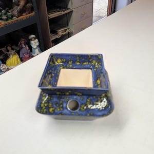 May include: A square ceramic oil warmer featuring a deep blue glaze with a yellow speckled pattern. The piece has a wide top basin for oil and a central circular opening on the side for a tea light candle, with a light cream interior finish.