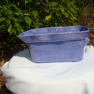 May include: A two-piece, rectangular ceramic baking dish set. The set is a speckled periwinkle blue color. The top piece has a slightly flared rim and handles. The set is sitting on a white cloth.
