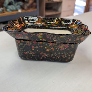 May include: A black ceramic dish with a rectangular base and a slightly flared, scalloped rim. The dish is decorated with colorful speckles of red, orange, yellow, and green. The dish is sitting on a white surface.