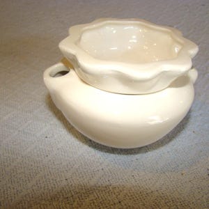 May include: A small, off-white ceramic container with a rounded base and a scalloped rim. The container has a small handle on one side and a matching lid. The piece is likely a decorative or functional item.