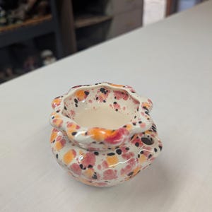 May include: A ceramic wax warmer featuring a scalloped rim and a rounded base. The piece is finished with a speckled glaze in shades of orange, pink, and black over a cream-colored background. A small circular opening is visible on the side.