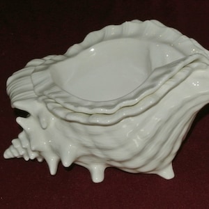 May include: A white ceramic seashell-shaped decorative bowl. The bowl has a textured, ridged exterior and a smooth, open interior. The bowl is supported by three small feet and is suitable for decorative purposes.