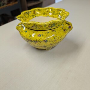 May include: A bright yellow ceramic planter with a speckled finish featuring blue and purple dots. The planter has a scalloped, ruffled rim and two small side handles, with a smooth, unglazed white interior basin visible at the top opening.