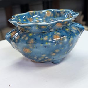 May include: A handcrafted ceramic bowl with a matching lid. The bowl and lid are a deep blue with speckled orange and yellow accents. The lid has a scalloped edge, and the bowl has three small feet.