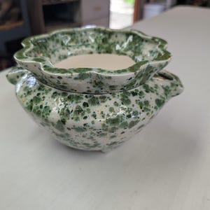 May include: A ceramic planter with a scalloped rim and two side handles, featuring a white base with a speckled green glaze pattern. The interior of the bowl is smooth and off-white, while the exterior displays a dense, mottled green finish.