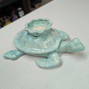May include: A light blue ceramic turtle figurine featuring a glossy, speckled glaze finish. The turtle has a scalloped, circular holder centered on its shell, designed to accommodate a small candle or tealight. The surface has a textured appearance.